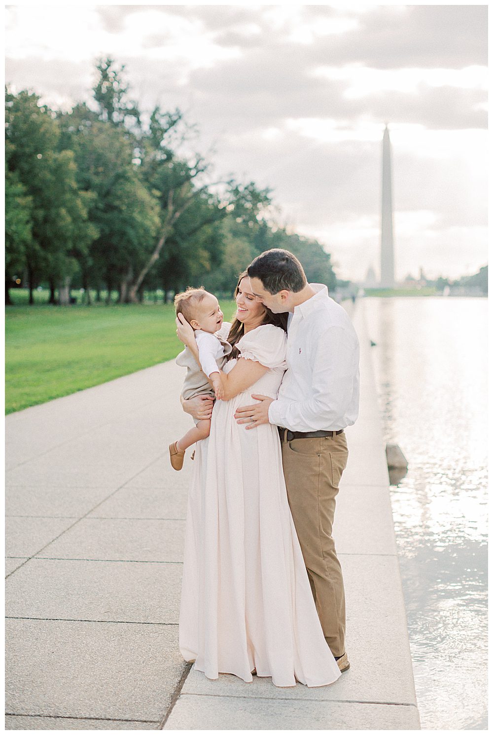 Parents hold their son and smile while standing in front of the reflecting pool in DC.