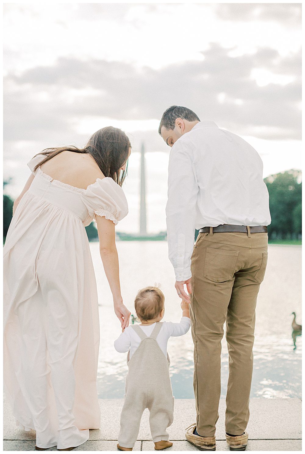 Parents lean down and hold toddler's hands while standing, looking out at DC reflecting pool during DC Monuments Family Photo Session.