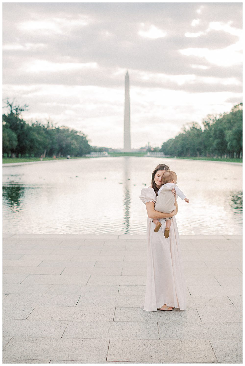 Mother holds her toddler up to cuddle while standing in front of Reflection Pool in DC.