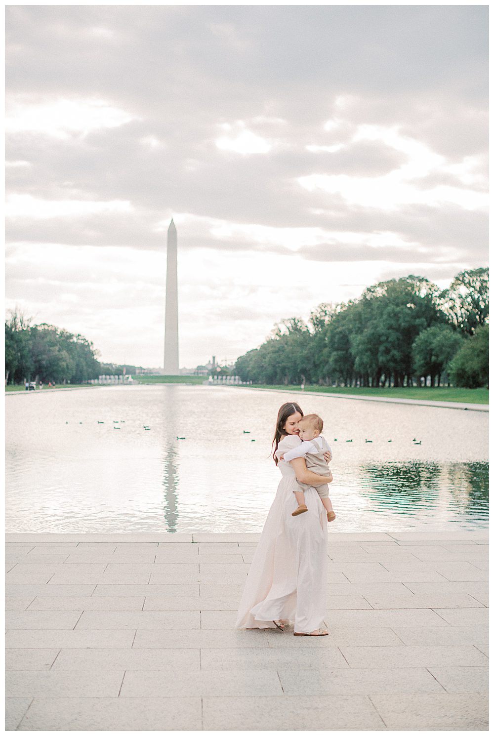 Mother walks in front of Washington monument while holding toddler son during DC Monuments Family Photo Session.