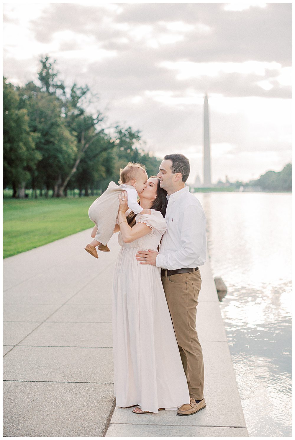 Mother holds up and kisses toddler's cheeks while standing with husband by DC Reflecting Pool.