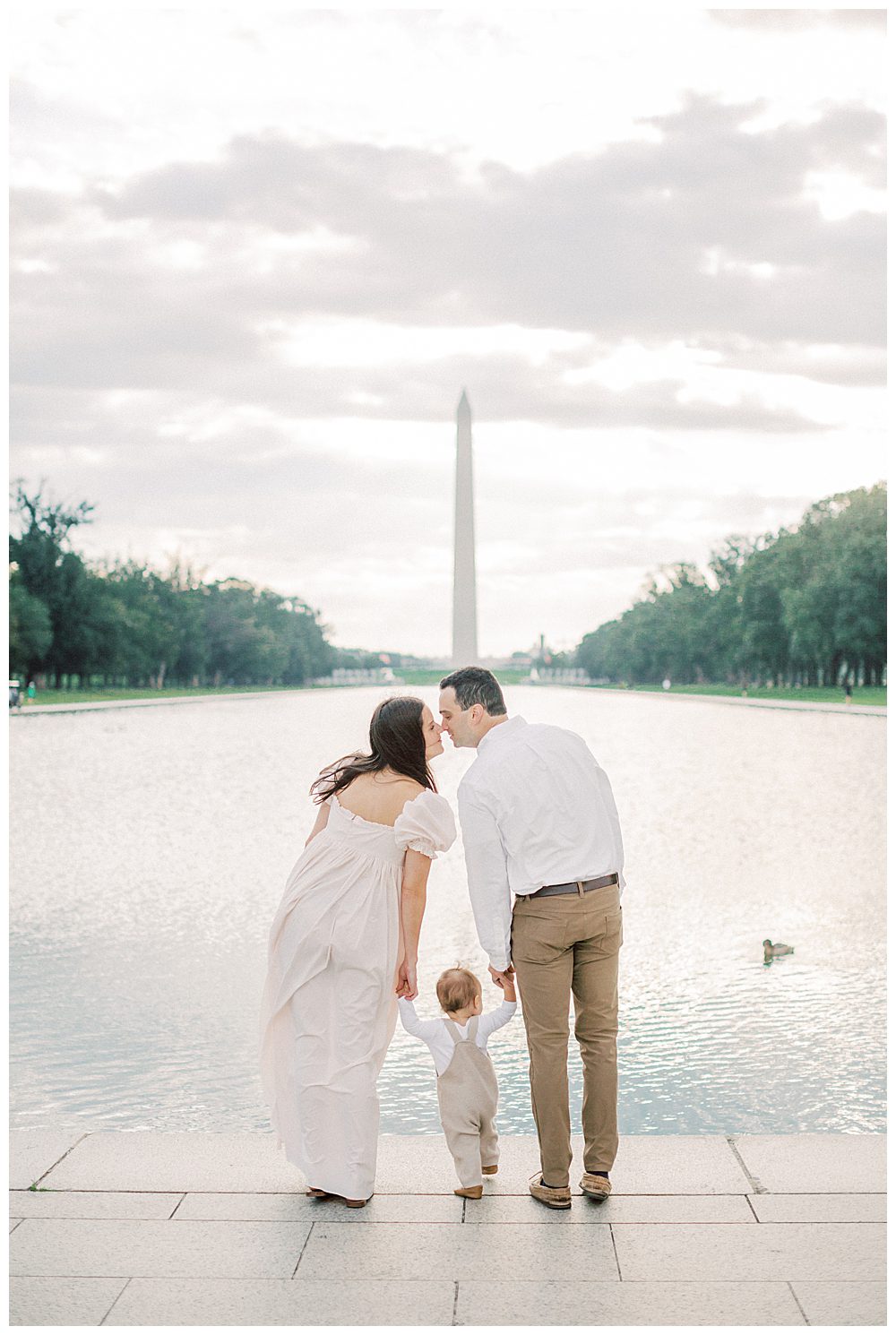 Mother and father lean in to kiss while holding their son's hand, standing in front of the Washington Monument and reflecting pool. 
