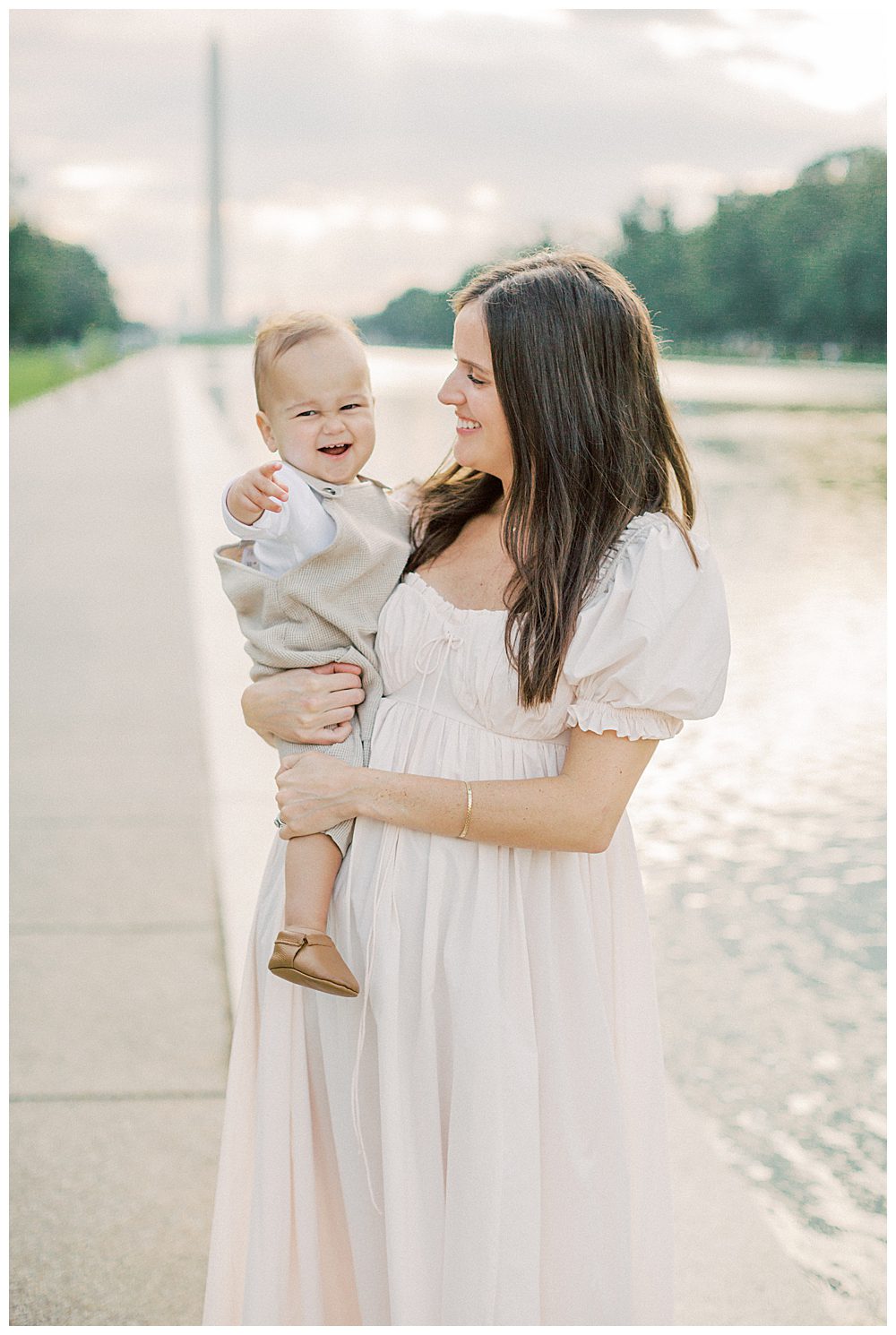 Mother smiles at her toddler son while holding him in front of the Reflecting Pool in Washington DC.