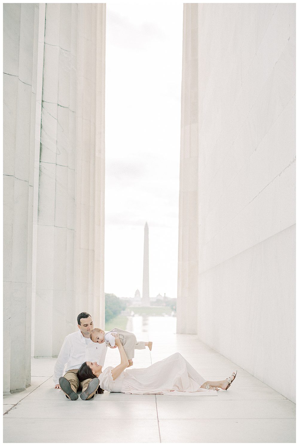 Mother holds up toddler son while laying in her husband's lap in the Lincoln Memorial during DC Monuments Family Photo Session.
