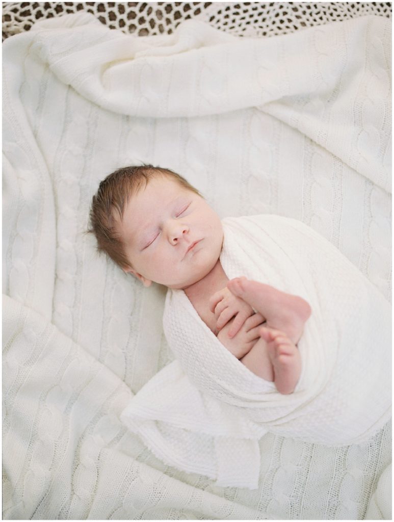 Baby girl with brown hair is swaddled on ivory knit blanket.