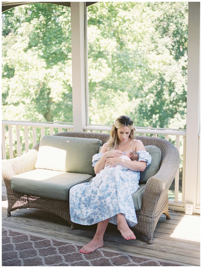 Mom sits on couch in sunroom, holding newborn baby girl.