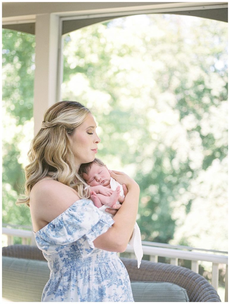 Blonde new mother holds newborn baby girl while standing in her sunroom during her Great Falls newborn session.