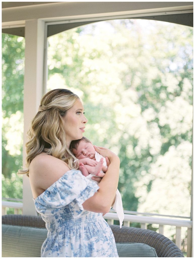 Blonde new mother holds newborn daughter in her sunroom, looking out her window.
