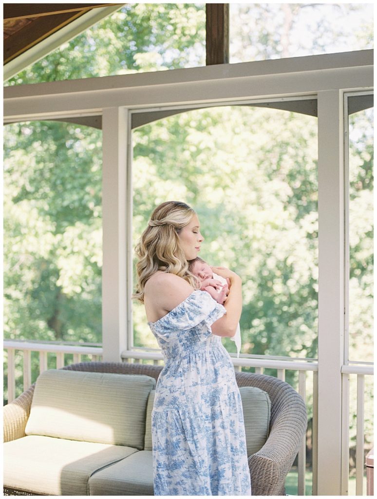 Blonde mother in blue Nothing Fits But dress holds baby girl in her sunroom and closes her eyes.