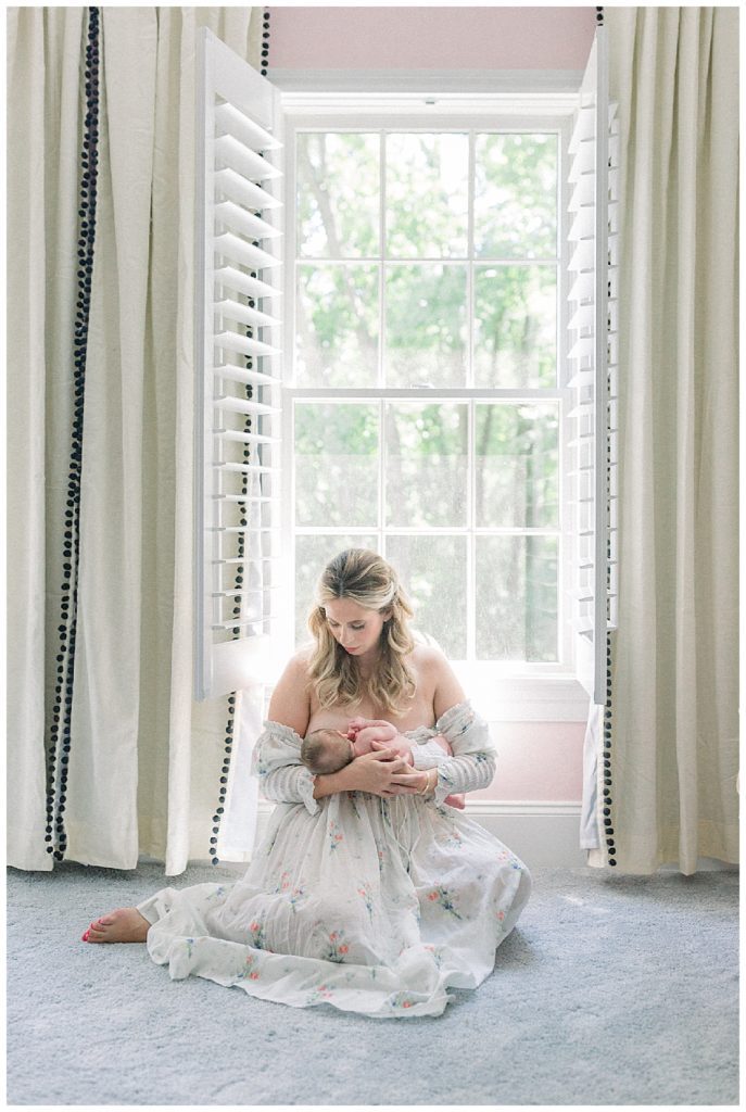 Blonde new mother nurses her baby girl while sitting in front of a large window, wearing the Doen Painted Floral dress.