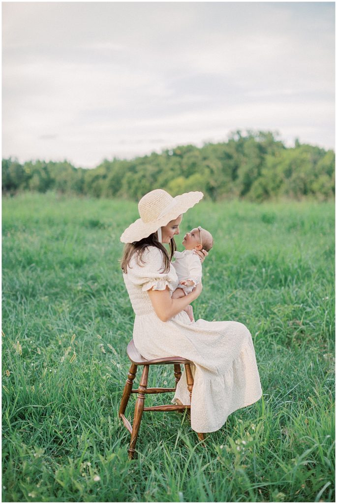 Mother holds newborn baby girl in front of her while sitting on a stool in a green field.