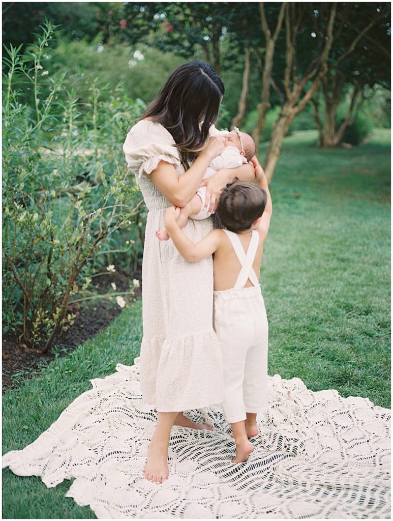 Toddler boy hugs mother as she holds newborn baby while they stand in a garden.