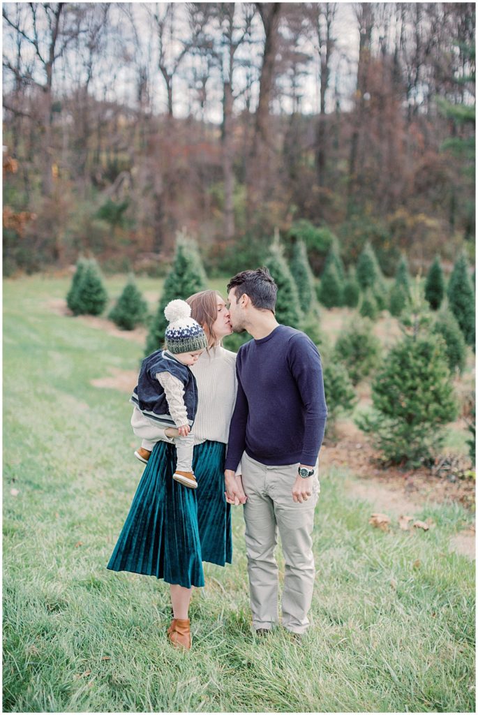 Parents hold their infant son and stop for a kiss at Christmas tree farm.