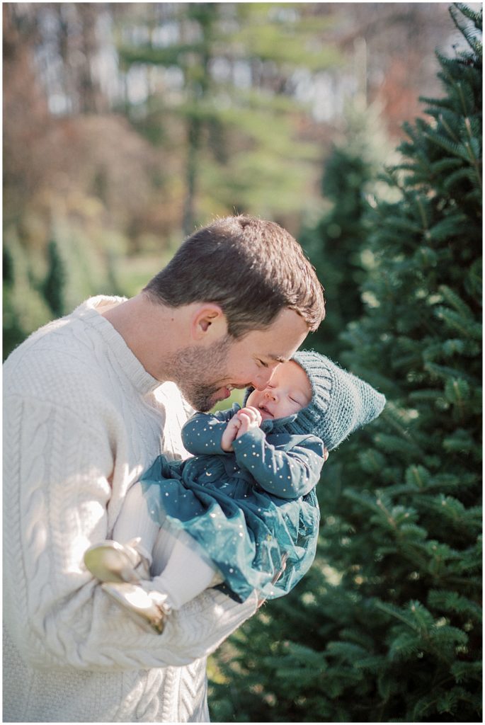 Father leans down and nuzzles his infant daughter's nose by a Christmas tree.