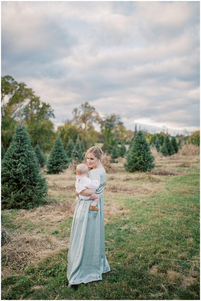 Mom in green satin dress holds baby during Christmas tree farm mini sessions at Butler's Orchard outside of Washington, DC.
