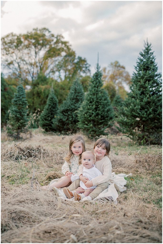 Three young children sit on blanket and smile during Christmas tree farm mini sessions at Butler's Orchard.