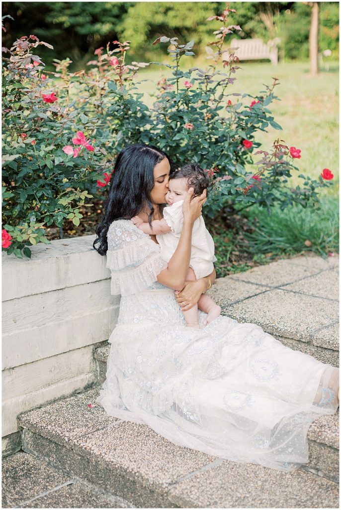 Mother holds infant daughter up close to her as she sits on steps in front of roses during Montgomery County Family Session at Brookside Gardens.