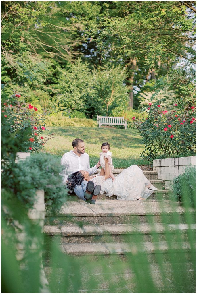 Mother, father, and infant daughter sit on steps surrounded by roses during Montgomery County Family Session at Brookside Gardens.