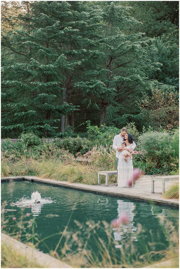 Mother and father stand by pond holding baby girl during Montgomery County Family Session at Brookside Gardens.