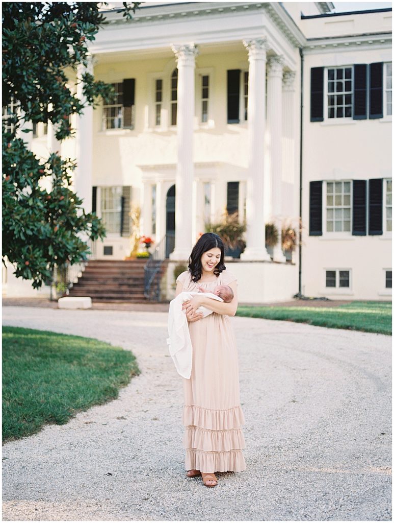 Mother smiles while holding her infant son in front of Oatlands Historic Home during newborn session by Loudoun County Newborn Photographer Marie Elizabeth Photography.