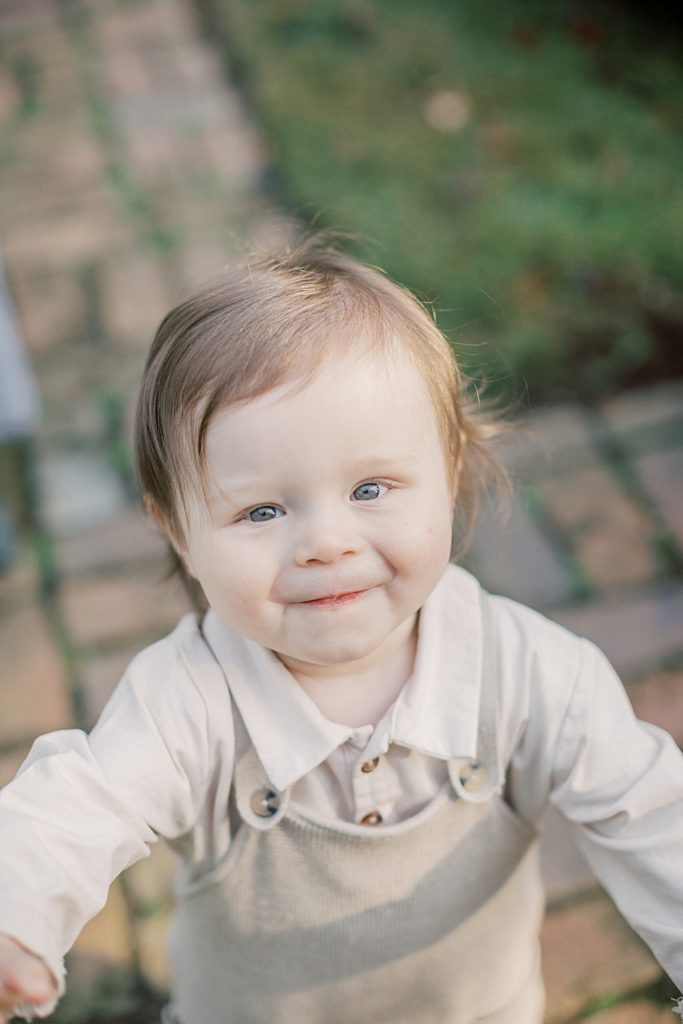 Baby boy in overalls with blue eyes smiles up at the camera photographed by Georgetown Family Photographer Marie Elizabeth Photography.