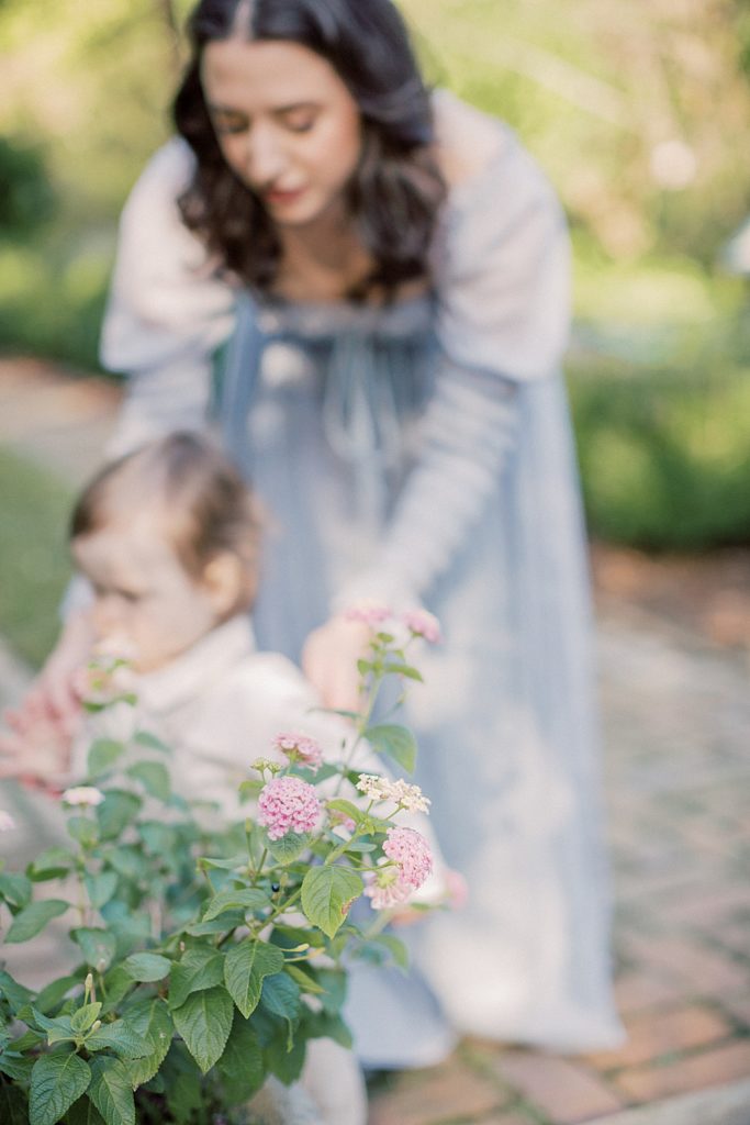 Mother in blue dress leans into help her toddler son walk in a garden photographed by Georgetown Family Photographer Marie Elizabeth Photography.