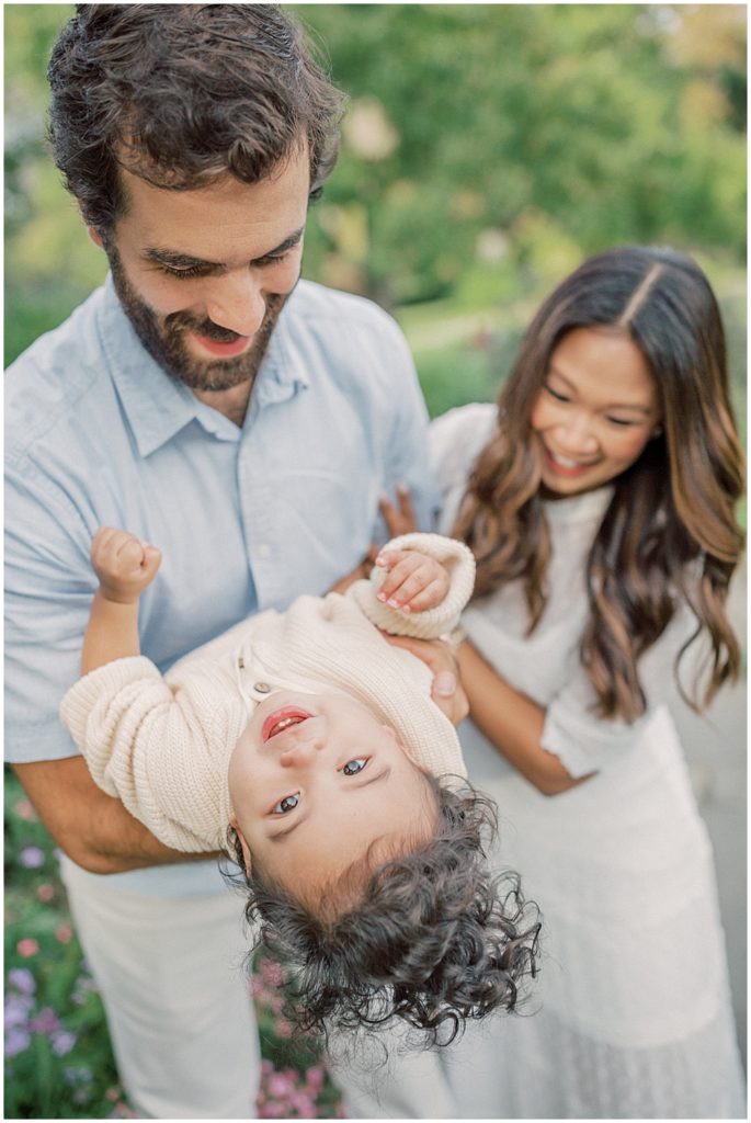 Baby boy with brown curly hair is held  outwards by parents, smiling during extended family session at Green Springs Gardens in Northern Virginia. 