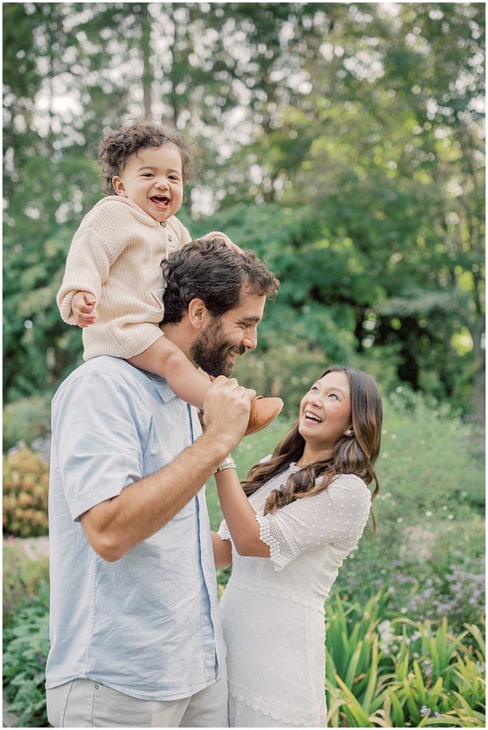 Parents laugh together as their toddler son sits on father's shoulders during extended family session at Green Springs Gardens in Northern Virginia. 