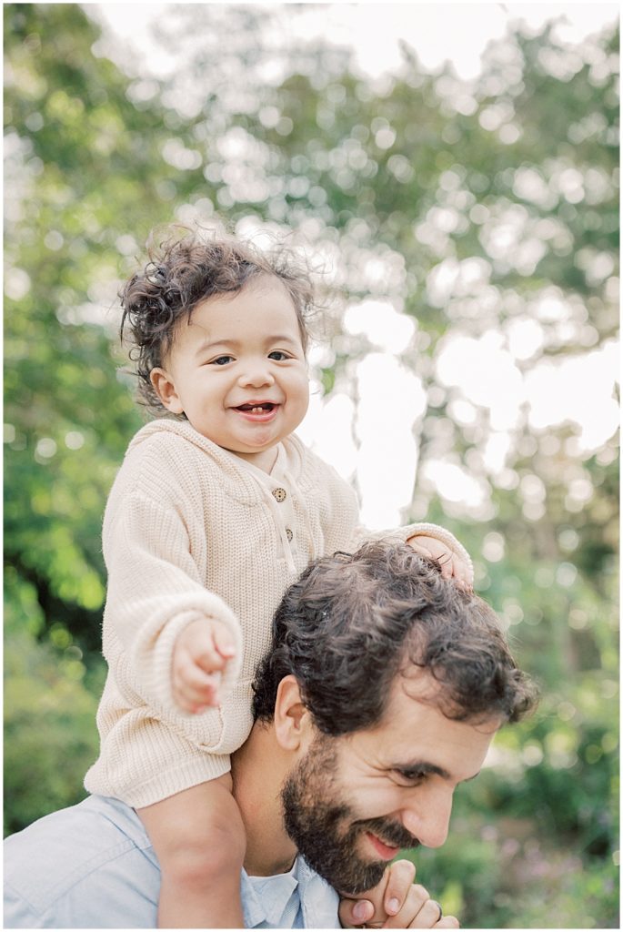Baby boy sits on dad's shoulders laughing during extended family session at Green Springs Gardens in Northern Virginia. 