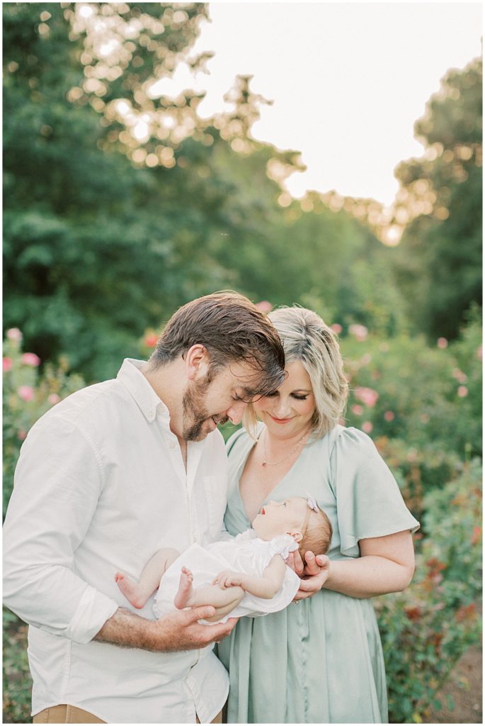 New parents smile down at baby girl during outdoor newborn family photos at Bon Air Rose Garden during sunset.