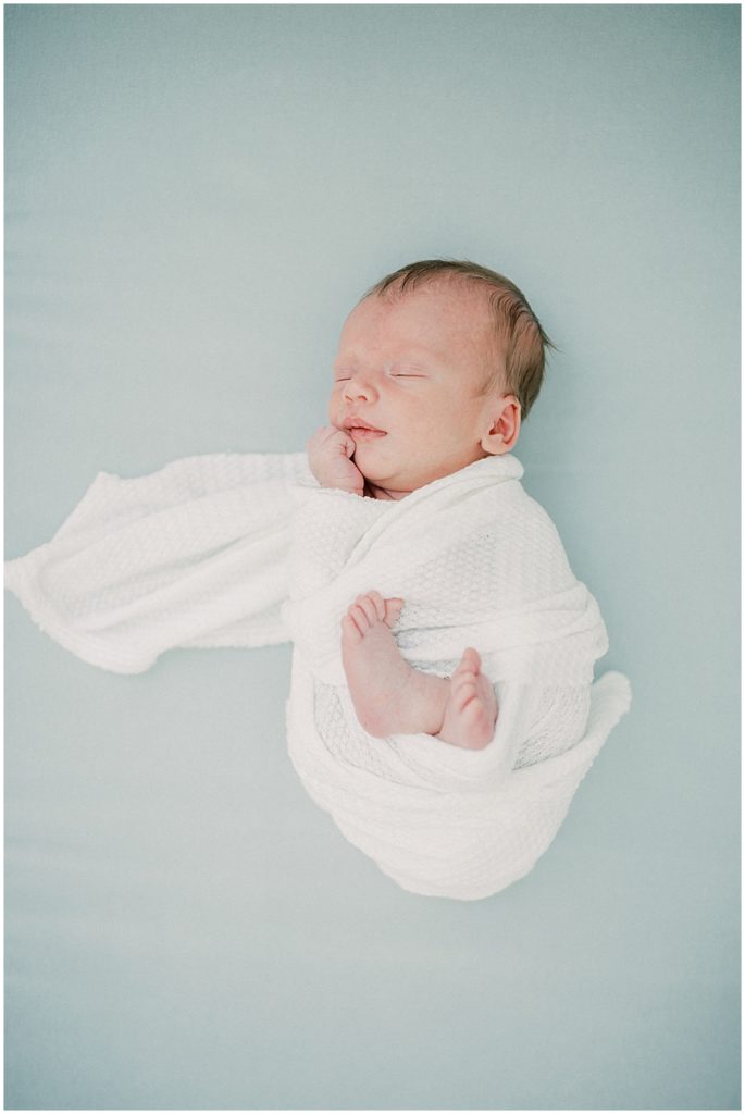 Baby boy swaddled in white sleeps on blue backdrop during studio newborn session.