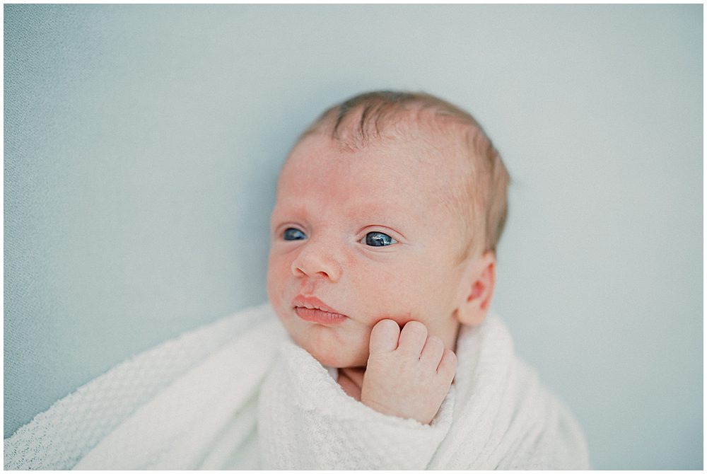 Baby boy with big blue eyes is swaddled in white and layed against a blue backdrop.