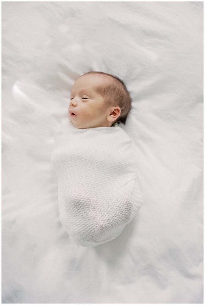 Newborn boy swaddle in white lays on white bed.
