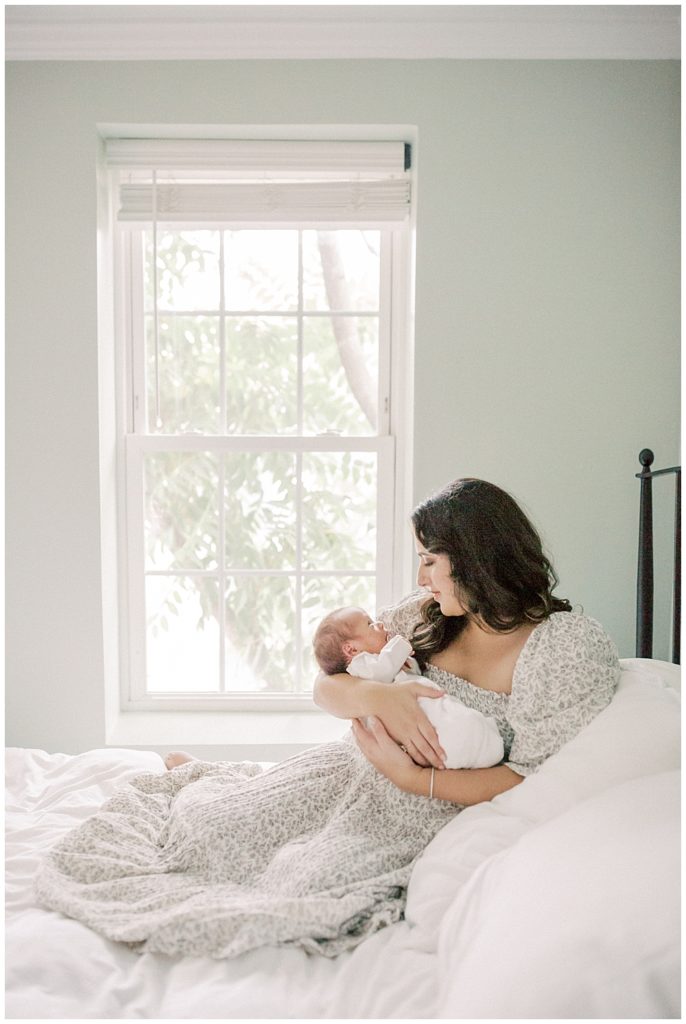 Mother sits on bed by window holding newborn during DC Row House Newborn Session.