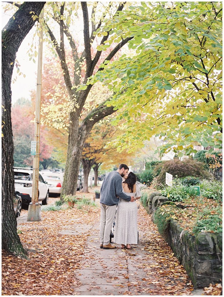 Man and woman stand on sidewalk and share a kiss on a fall day.