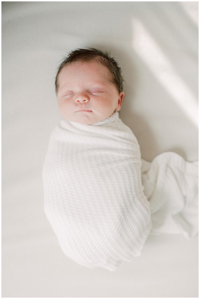 Baby girl swaddled in white lays on an ivory mat.