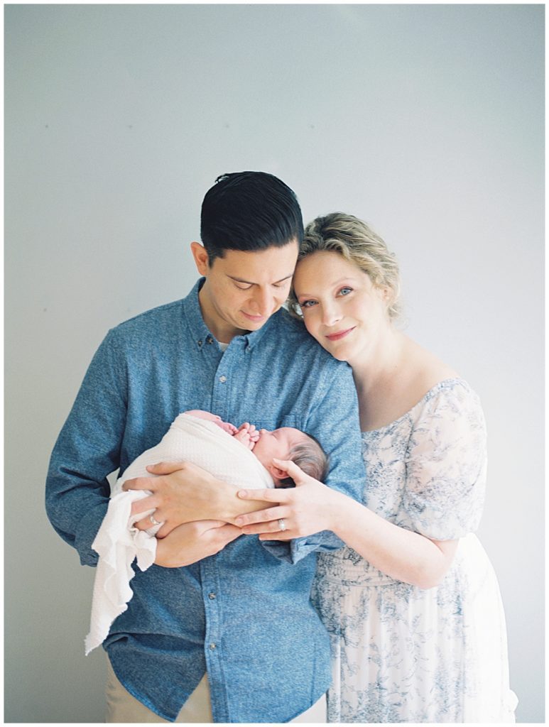 Mother with blonde hair gives soft smile at the camera while leaning on her husband during Fairfax VA newborn session.