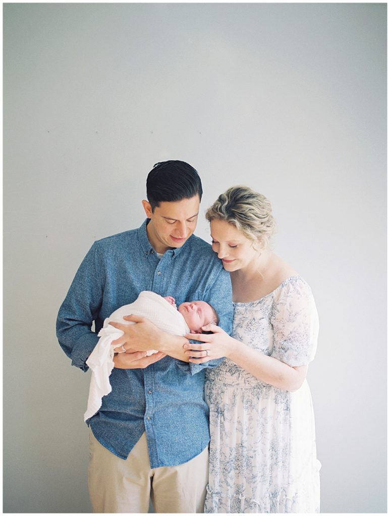 New parents stand together and smile at newborn baby girl in their home during Fairfax VA newborn session.