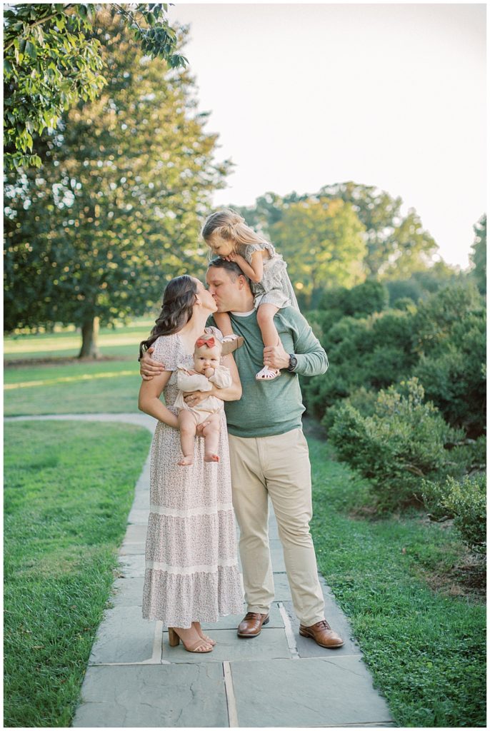 Mother and father hold their two daughter while leaning in for a kiss during Glenview Mansion Photos by Marie Elizabeth Photography.
