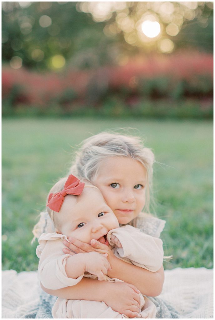 Toddler girl holds her baby sister on a blanket in the grass.