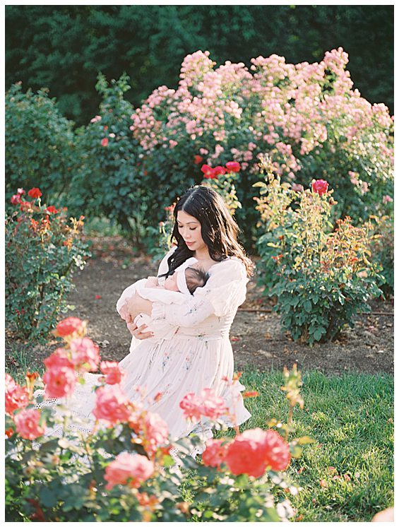 Dark-haired mother kneels in Bon Air Rose Garden holding her baby girl photographed by Alexandria Newborn Photographer Marie Elizabeth Photography.