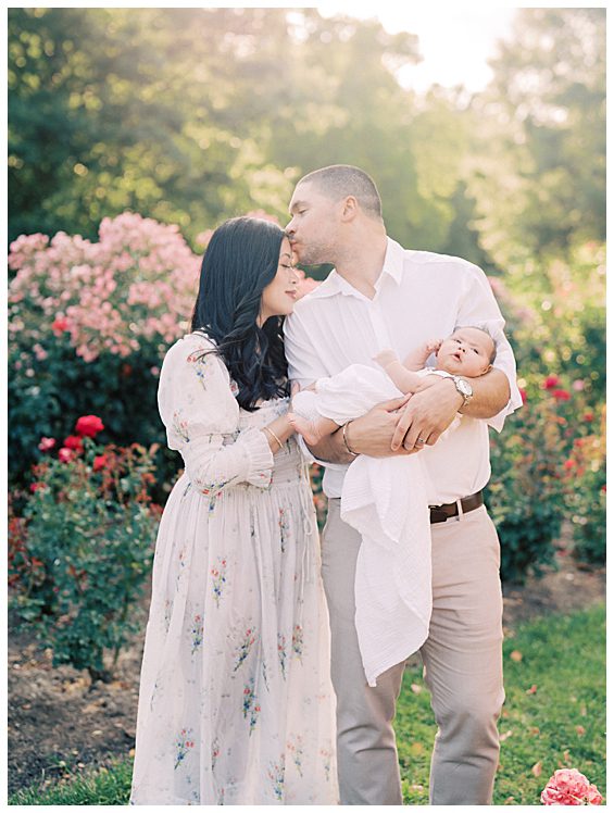 Father leans over and kisses his wife's forehead while holding baby girl photographed by Alexandria Newborn Photographer Marie Elizabeth Photography.