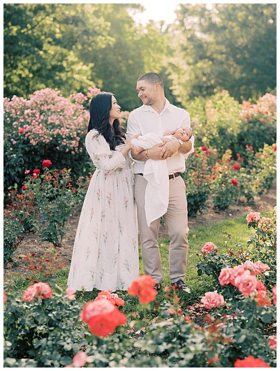 New parents hold their baby girl while standing in a rose garden photographed by Alexandria Newborn Photographer Marie Elizabeth Photography.