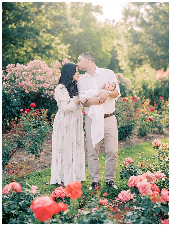 Father holds newborn daughter and kisses his dark-haired wife's forehead in Bon Air Rose Garden photographed by Alexandria Newborn Photographer Marie Elizabeth Photography.