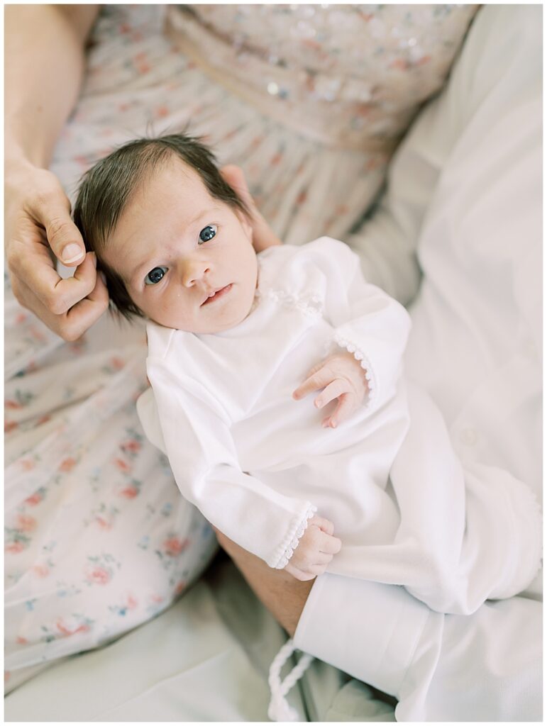 Baby girl, held by her parents, looks up at the camera with blue eyes photographed by Arlington Newborn Photographer Marie Elizabeth Photography.