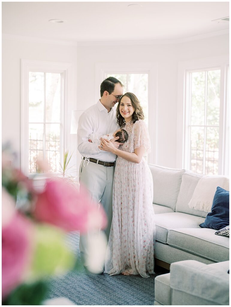A mother in a Needle & Thread dress smiles at the camera as her husband holds their baby girl and leans into her while standing in their home, photographed by Arlington Newborn Photographer Marie Elizabeth Photography.