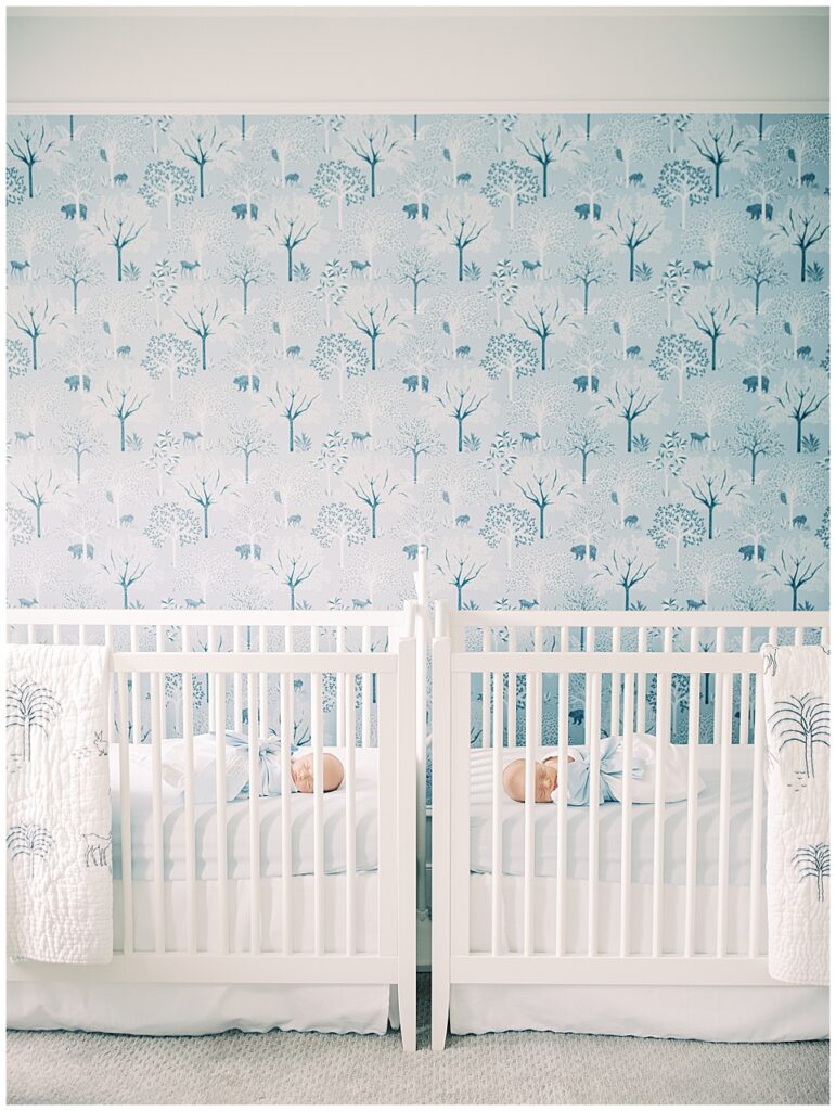 Twin baby boys sleep in their cribs in a blue wallpaper Serena & Lily nursery.