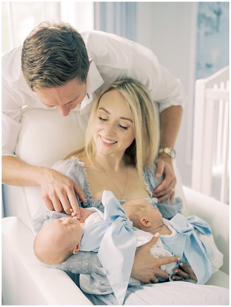 Blonde father leans over chair where his wife holds their twin baby boys and grasps the hand of the one of the babies.