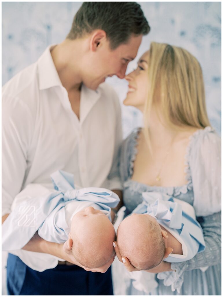Mother and father lean into one another and smile as they hold their twin baby boys in Beaufort Bonnet bow swaddles in front of them.