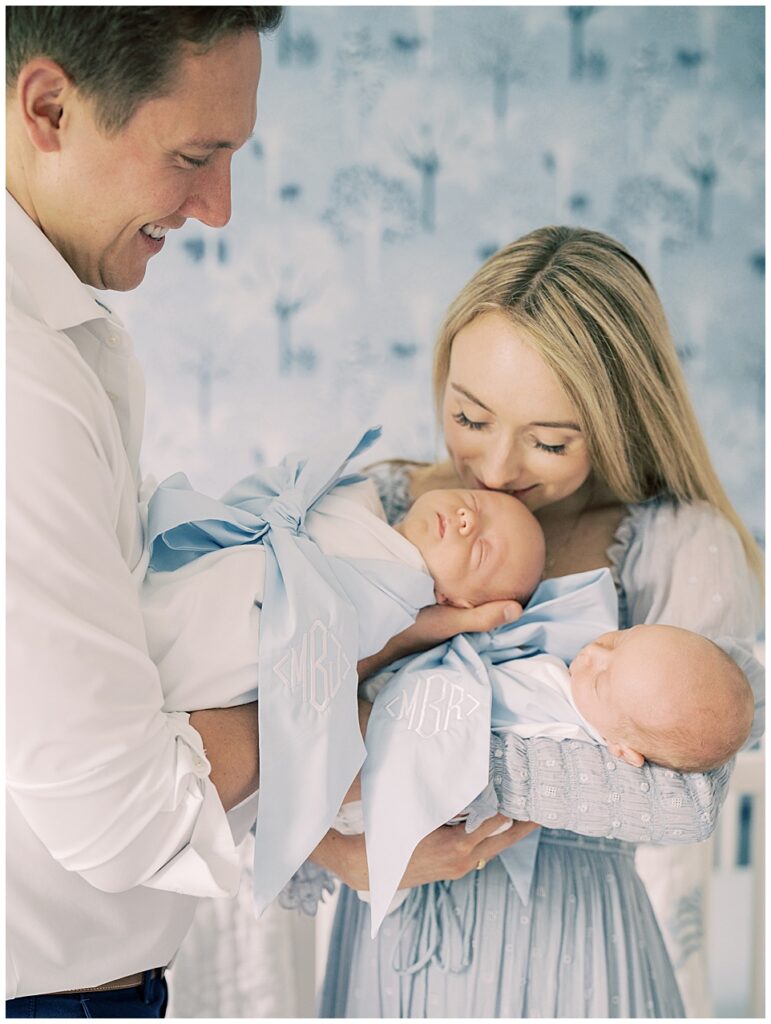Mother leans down to kiss one of her twin babies wearing a Beaufort Bonnet bow swaddle as her husband holds him while she holds the other baby in their Serena & Lily nursery.